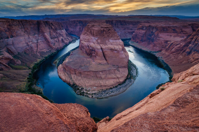 This landscape photograph captures the breathtaking view of Horseshoe Bend, a stunning natural formation on the Colorado River near Page, Arizona. The image showcases the iconic, horseshoe-shaped meander carved by the river, surrounded by towering red rock cliffs. The rich, vibrant colors of the rocks contrast with the deep turquoise waters of the river, which glimmers under the bright desert sunlight. The vast, open sky above adds to the sense of scale and majesty, while subtle shadows cast by the cliffs enhance the texture and depth of the scene. The photograph captures the dramatic beauty and grandeur of this iconic Southwest landmark.