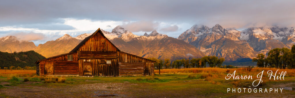 The TA Moulton barn is a classic wooden barn with a weathered, rustic appearance sits in the foreground of the panorama image. The barn, with its slanted gable roof, and wooden siding, is framed by the expansive, open fields of Mormon Row. In the background, the majestic Teton Range rises dramatically against a cloudy sky above. The lighting is early morning, casting a warm, golden hue over the scene. The barn’s structure and the surrounding landscape reflect a serene and picturesque rural setting.