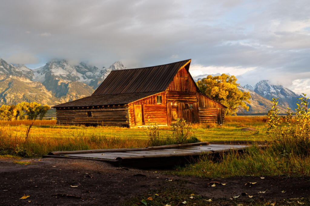 Scenic view of TA Moulton Barn, a historic wooden barn with a classic, rustic charm, set against a backdrop of the majestic Teton Range. In the foreground, a charming walking bridge spans a gentle stream, enhancing the picturesque rural landscape. The barn, with its iconic gabled roof and weathered wood, contrasts beautifully with the surrounding lush greenery and the towering mountain peaks in the distance.