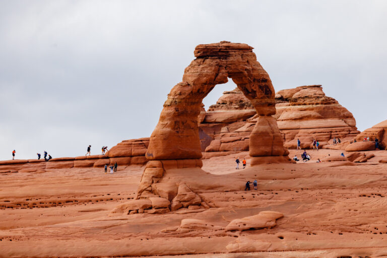 Delicate Arch in Arches National Park with hikers in the foreground, highlighting the scale and grandeur of the iconic sandstone formation under a cloudy sky.