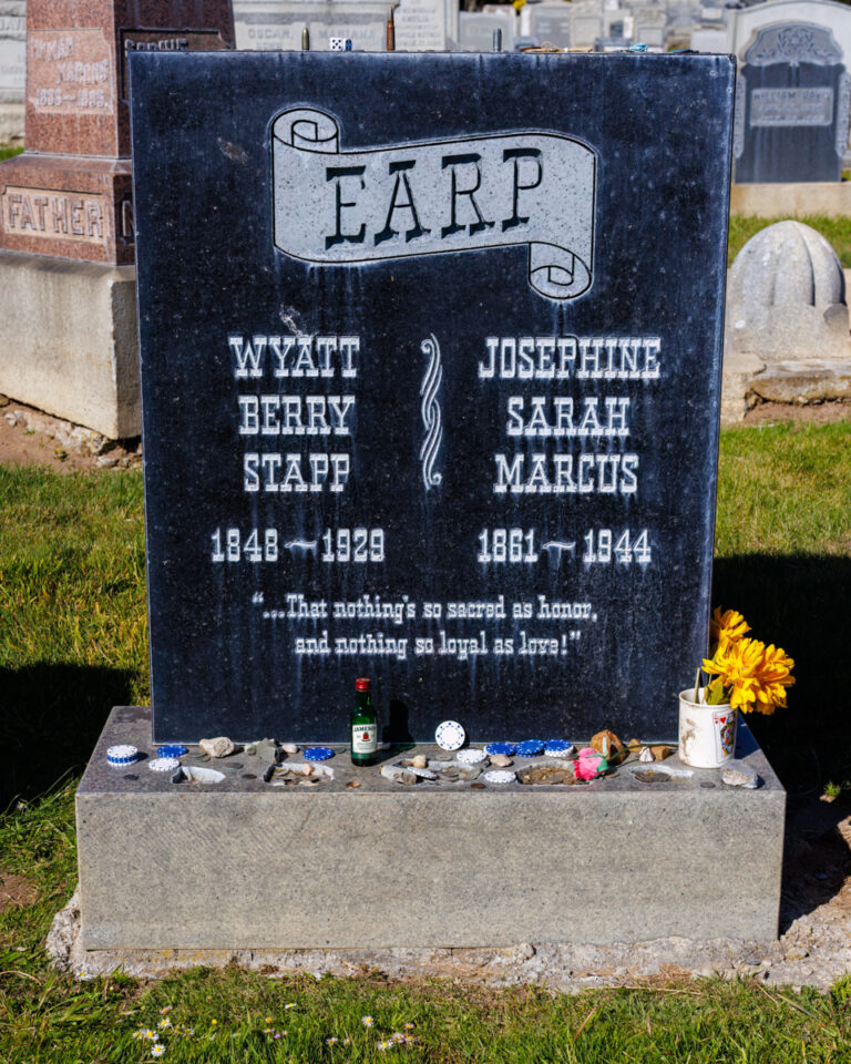 Headstone of Wyatt Berry Stapp Earp and Josephine Sarah Marcus Earp at a cemetery, adorned with personal tributes like flowers, poker chips, and a small bottle of whiskey, with other gravestones visible in the background