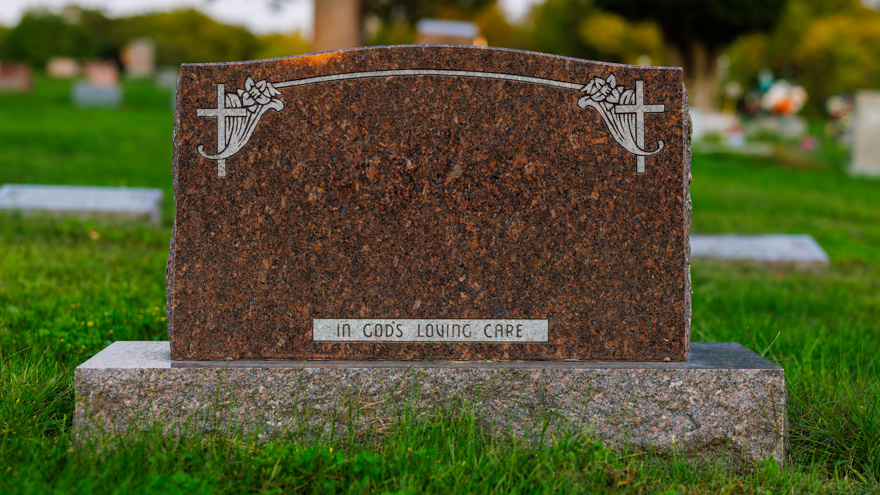 Blank headstone engraved with the inscription "In God's Loving Care" at the bottom, set in a grassy cemetery with other blurred headstones in the background.