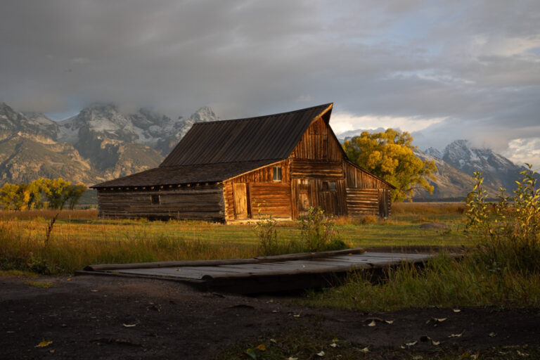 RAW / unedited photo of a scenic view of TA Moulton Barn, a historic wooden barn with a classic, rustic charm, set against a backdrop of the majestic Teton Range. In the foreground, a charming walking bridge spans a gentle stream, enhancing the picturesque rural landscape. The barn, with its iconic gabled roof and weathered wood, contrasts beautifully with the surrounding lush greenery and the towering mountain peaks in the distance.